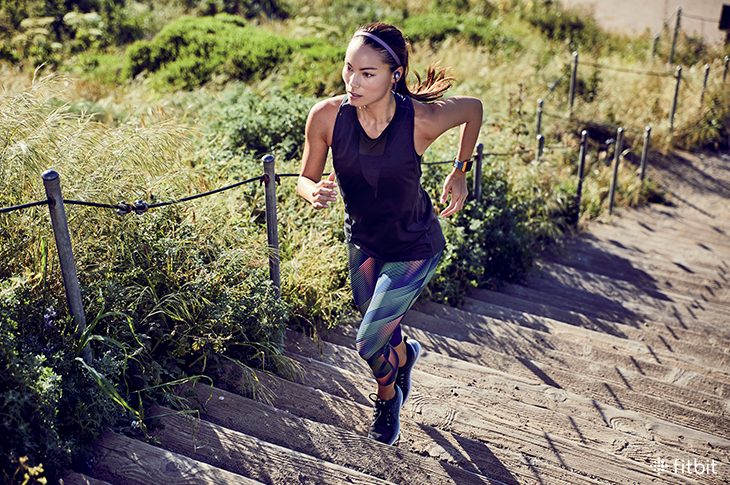 Young woman, maybe asian, wearing workout clothes and running up stairs in a park or other pretty outdoor location. She's wearing flyer in nightfall blue and ionic in slate blue and burnt orange.